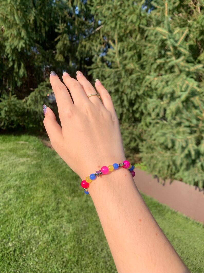 Pansexual pride flag gemstone bracelet being worn on someone's wrist. There are green bushes in the background and it is sunny.