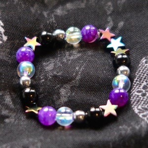 Puede incluir: Un brazalete de cuentas de color morado, negro y plateado con cuentas iridiscentes y abalorios de estrellas doradas.