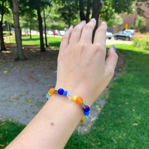 Aroace pride flag gemstone bracelet being worn on someone's wrist. There are green bushes in the background and it is sunny.