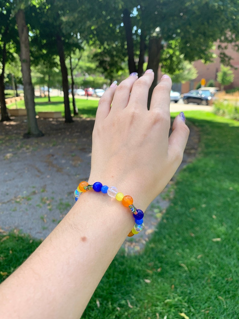 Aroace pride flag gemstone bracelet being worn on someone's wrist. There are green bushes in the background and it is sunny.