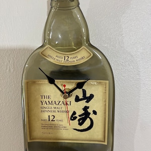 Puede incluir: Una botella de vidrio transparente con una tapa dorada, con forma de botella de sake japonesa tradicional. La botella tiene una etiqueta con el texto "The Yamazaki Single Malt Japanese Whisky Aged 12 Years" y un carácter japonés en tinta negra. La botella es un reloj con manecillas negras y un centro dorado.