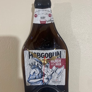 May include: A brown glass bottle-shaped clock with the Hobgoblin Ruby Beer label. The clock features gold-colored hands and the text "Hobgoblin Ruby Beer". The clock is mounted on a wall.