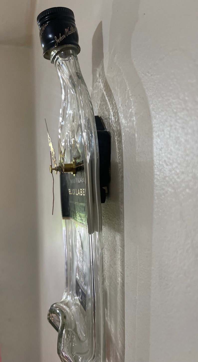 Puede incluir: Una botella de vidrio transparente con una etiqueta negra que dice "Johnnie Walker Black Label" se ha reutilizado como reloj de pared. Las manecillas del reloj son doradas y el mecanismo del reloj est&aacute; unido a la parte posterior de la botella.