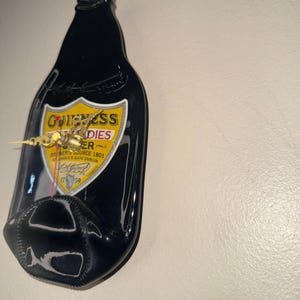 Puede incluir: Un reloj de botella de cerveza Guinness negra con una etiqueta amarilla y blanca. Las manecillas del reloj son doradas. La etiqueta dice "Guinness Foreign Extra Stout". El reloj est&aacute; montado en una pared blanca.