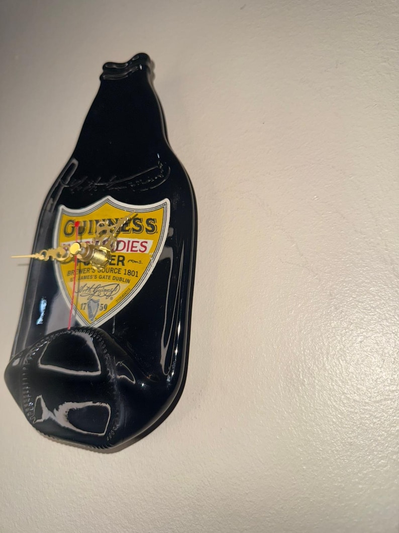 Puede incluir: Un reloj de botella de cerveza Guinness negra con una etiqueta amarilla y blanca. Las manecillas del reloj son doradas. La etiqueta dice "Guinness Foreign Extra Stout". El reloj est&aacute; montado en una pared blanca.