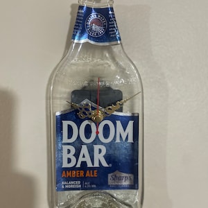 May include: A clear glass beer bottle with a clock face and hands. The bottle has a blue label with the text "Doom Bar Amber Ale" and "Sharp's Brewery".