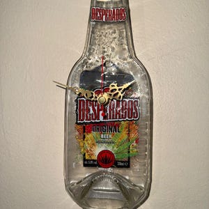 Può includere: Un orologio ricavato da una bottiglia di birra Desperados riciclata. Il quadrante dell'orologio è visibile attraverso il vetro, con lancette color oro e un meccanismo centrale. La bottiglia ha testo rosso e nero.