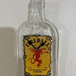 Puede incluir: Una botella de vidrio transparente con una etiqueta de licor de whisky de canela Fireball, reutilizada como reloj. La etiqueta presenta un demonio rojo, caliente y ardiente con las palabras "Fireball", "Red" y "Hot". La esfera del reloj es negra con manecillas blancas.