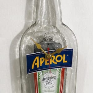 May include: A repurposed glass bottle clock with a blue cap and gold clock hands. The label reads "APEROL APERITIVO 1919" in white and yellow against a red and green background. The clock is a unique home decor item.