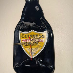 Puede incluir: Un reloj de pared negro con forma de botella de Guinness. La esfera del reloj presenta una etiqueta amarilla y blanca con el logotipo y el texto de Guinness. Las manecillas del reloj son doradas y el segundero es rojo.