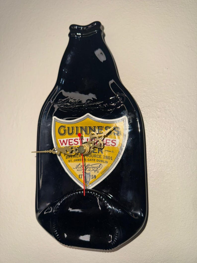 Puede incluir: Un reloj de pared negro con forma de botella de Guinness. La esfera del reloj presenta una etiqueta amarilla y blanca con el logotipo y el texto de Guinness. Las manecillas del reloj son doradas y el segundero es rojo.