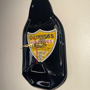 Puede incluir: Un reloj de pared negro con forma de botella de Guinness. La esfera del reloj presenta el logotipo de Guinness con manecillas doradas y el texto "BREWER'S SOURCE 1801 ST. JAMES'S GATE DUBLIN". El reloj est&aacute; montado en una pared blanca.