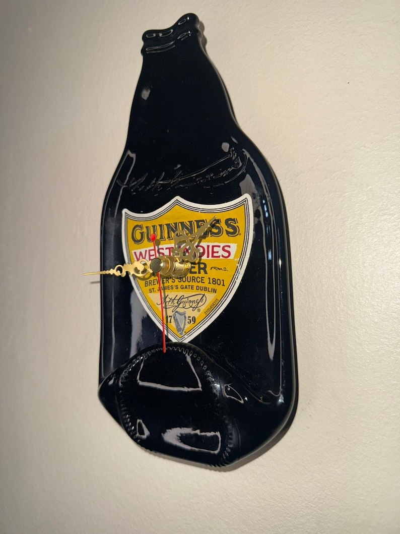 Puede incluir: Un reloj de pared negro con forma de botella de Guinness. La esfera del reloj presenta el logotipo de Guinness con manecillas doradas y el texto "BREWER'S SOURCE 1801 ST. JAMES'S GATE DUBLIN". El reloj est&aacute; montado en una pared blanca.