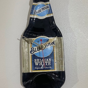 Puede incluir: Un reloj de botella de vidrio azul con forma de botella de cerveza Blue Moon Belgian White. La esfera del reloj es blanca con detalles azules y rojos. La botella tiene una etiqueta que dice "Blue Moon Brewing Company" y "Elaborada con cáscara de naranja para un refrescante sabor cítrico".