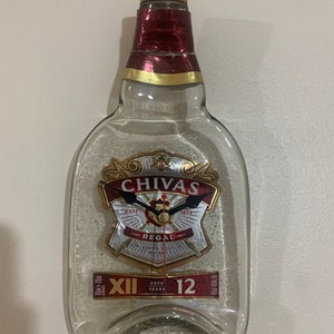 May include: A clear glass bottle with a red and gold label that reads "Chivas Regal Blended Scotch Whisky" and "Aged 12 Years". The bottle has been repurposed into a clock with black hands and Roman numeral markings.