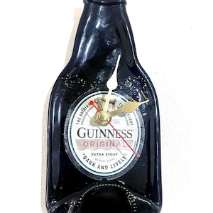 Puede incluir: Un reloj de botella de cerveza Guinness negra con una esfera dorada y agujas rojas. La esfera del reloj tiene el logotipo de Guinness y el texto "The Original Est. 1778 X Stout Guinness Original Extra Stout Original Recipe "Dark and Lively".