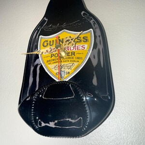 Puede incluir: Un reloj de pared negro con forma de botella de Guinness. La esfera del reloj presenta el logotipo de Guinness en amarillo y negro, con las palabras "PORTER" y "ST. JAMES'S GATE DUBLIN". Las manecillas del reloj son doradas.