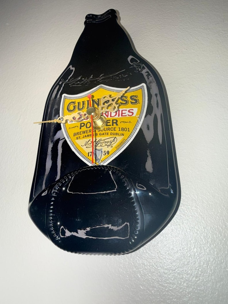 Puede incluir: Un reloj de pared negro con forma de botella de Guinness. La esfera del reloj presenta el logotipo de Guinness en amarillo y negro, con las palabras "PORTER" y "ST. JAMES'S GATE DUBLIN". Las manecillas del reloj son doradas.