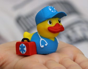 May include: A yellow rubber duck wearing a blue medical scrubs outfit with a red first aid kit and a stethoscope. The duck is wearing a blue hat with a white star of life.