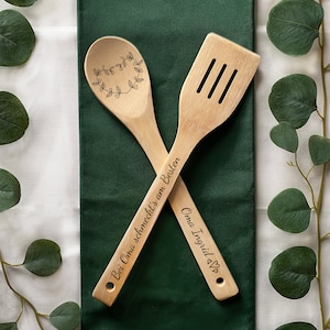 May include: Two wooden kitchen utensils, a spoon and a spatula, with engraved text. The spoon has a heart-shaped wreath design and the text "Bei Oma schmeckt's am Besten". The spatula has the text "Oma Ingrid &hearts;".