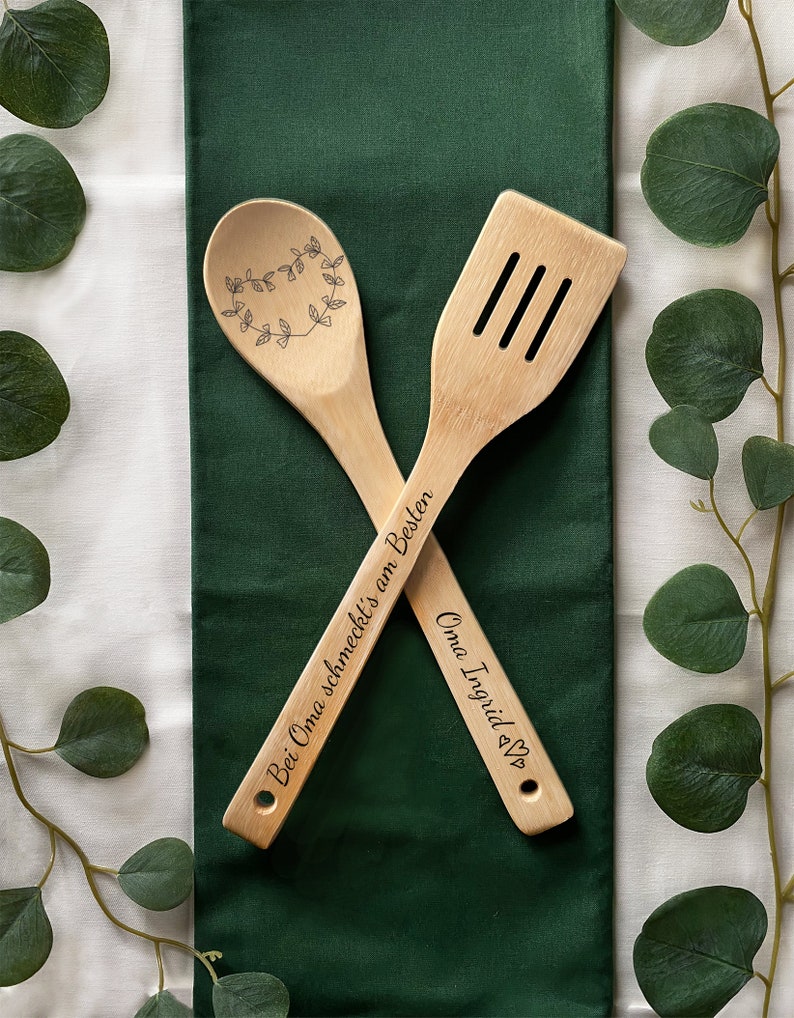 May include: Two wooden kitchen utensils, a spoon and a spatula, with engraved text. The spoon has a heart-shaped wreath design and the text "Bei Oma schmeckt's am Besten". The spatula has the text "Oma Ingrid &hearts;".