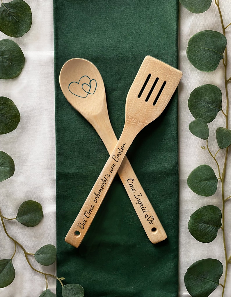 May include: Two wooden kitchen utensils with engraved text. The larger utensil is a spatula with the text "Oma Ingrid &hearts;". The smaller utensil is a spoon with the text "Bei Oma schmeckt's am Besten".