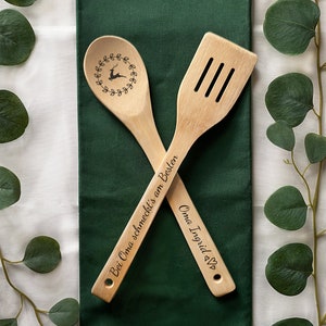 May include: Two wooden kitchen utensils with engraved text. The spoon has a reindeer design and the text "Bei Oma schmeckt's am Besten". The spatula has the text "Oma Ingrid &hearts;".