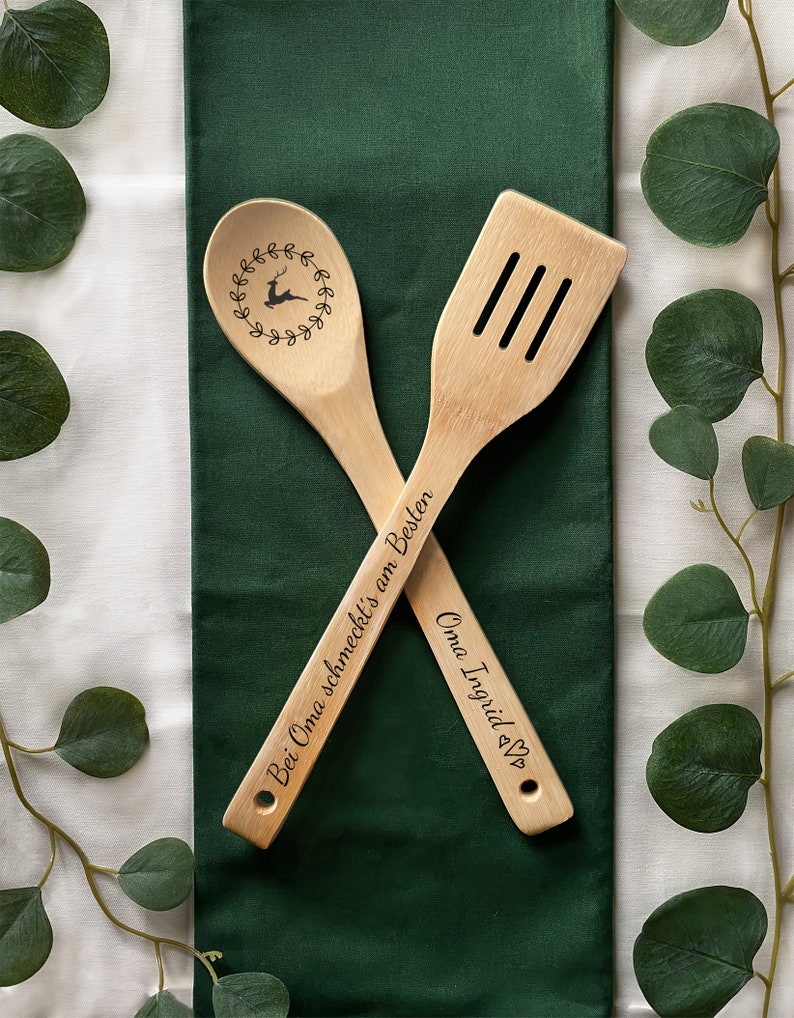 May include: Two wooden kitchen utensils with engraved text. The spoon has a reindeer design and the text "Bei Oma schmeckt's am Besten". The spatula has the text "Oma Ingrid &hearts;".