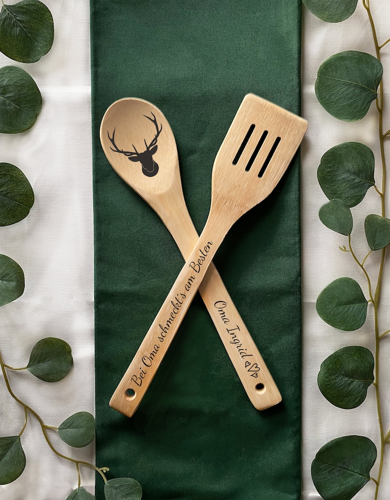 May include: Two wooden kitchen utensils, a spoon and a spatula, with engraved text. The spoon has a deer silhouette and the text "Bei Oma schmeckt's am Besten". The spatula has the text "Oma Ingrid <3".