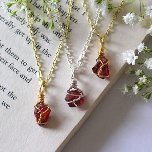 May include: Three necklaces featuring raw, red gemstone pendants. The gemstones are wrapped in gold and silver wire and suspended from delicate chains. The necklaces are displayed on a book page, with white flowers.