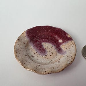 Può includere: Una piccola ciotola rotonda in ceramica con una base screziata bianco sporco e una glassa marrone che cola sul lato. Una moneta è accanto alla ciotola. La ciotola è probabilmente fatta a mano.