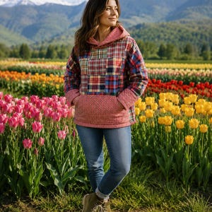 May include: A patchwork hoodie with a red, blue, and green plaid pattern, featuring a red hood and pocket, paired with blue jeans and olive-green boots. The background shows a field of colorful tulips and mountains.