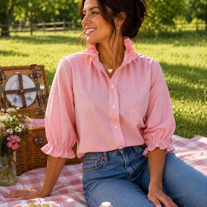Può includere: Una camicia abbottonata a quadretti vichy rosa e bianchi con colletto a volant e maniche a tre quarti con polsini arricciati. La camicia è abbinata a jeans blu e a una collana d'oro con ciondolo a forma di cuore.