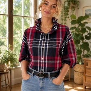 May include: A button-down shirt featuring a red, navy blue, and white plaid pattern with a ruffled collar, three-quarter length sleeves, and a small chest pocket. The shirt is paired with light blue denim jeans and a black leather belt.