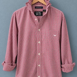 Puede incluir: Una camisa de manga larga con botones en un patrón de cuadros vichy rojo y blanco. La camisa presenta un cuello con volantes, una tapeta de botones frontal y un bolsillo en el pecho con un pequeño logotipo bordado en blanco. La etiqueta interior dice Nobby Clothes Shop.