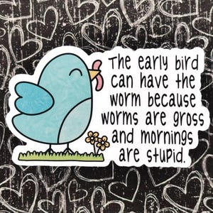 Op de afbeelding: Een gestanste sticker met een lichtblauwe cartoonvogel die een roze worm in zijn snavel houdt, staand op groen gras naast twee kleine gele bloemen. De tekst luidt: The early bird can have the worm because worms are gross and mornings are stupid.