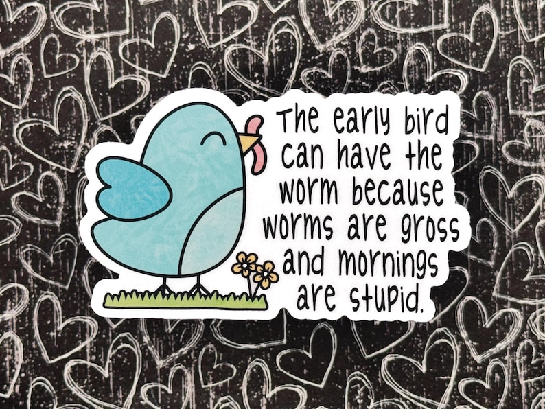 Op de afbeelding: Een gestanste sticker met een lichtblauwe cartoonvogel die een roze worm in zijn snavel houdt, staand op groen gras naast twee kleine gele bloemen. De tekst luidt: The early bird can have the worm because worms are gross and mornings are stupid.