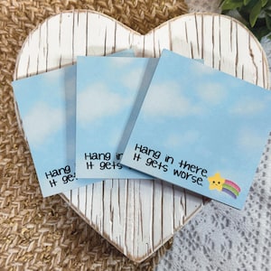 May include: Three square sticky notes featuring a light blue sky with white clouds. Each note displays the black text "Hang in there. It gets worse." and a small yellow star graphic with a colorful rainbow trail in the bottom right corner.