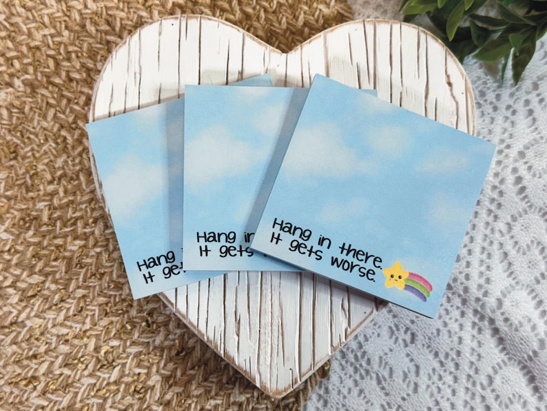 May include: Three square sticky notes featuring a light blue sky with white clouds. Each note displays the black text "Hang in there. It gets worse." and a small yellow star graphic with a colorful rainbow trail in the bottom right corner.