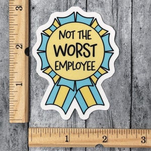 May include: A sticker in the shape of a ribbon with the text "NOT THE WORST EMPLOYEE". The ribbon has a yellow center with blue and yellow accents. The sticker is on a gray wooden surface.