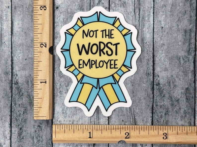 May include: A sticker in the shape of a ribbon with the text "NOT THE WORST EMPLOYEE". The ribbon has a yellow center with blue and yellow accents. The sticker is on a gray wooden surface.