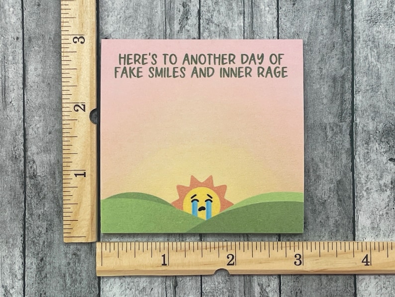 Pu&ograve; includere: Un tramonto rosa e arancione con un viso di sole giallo che piange su colline verdi. Il testo dice "HERE'S TO ANOTHER DAY OF FAKE SMILES AND INNER RAGE".
