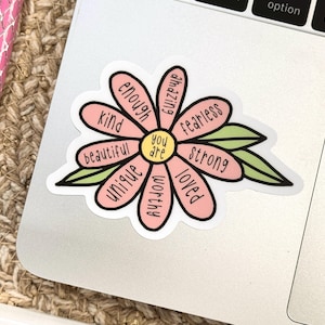 Peut inclure: Un autocollant en forme de fleur avec des p&eacute;tales roses, chacun portant un mot imprim&eacute; en noir : enough, amazing, fearless, strong, loved, worthy, unique, beautiful et kind. Le centre est jaune avec le texte you are et deux feuilles vertes.