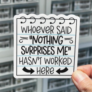 May include: A square white sticker with a scalloped top edge resembling a notepad page. It features black text that reads: WHOEVER SAID "NOTHING SURPRISES ME" HASN'T WORKED HERE, accented by thin blue horizontal lines and two black arrows pointing inward.