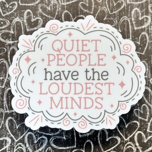 May include: A die-cut sticker with a white background featuring the text "QUIET PEOPLE have the LOUDEST MINDS" in pink and gray lettering. The design is enclosed in a decorative gray and pink scalloped border with small star and swirl accents.