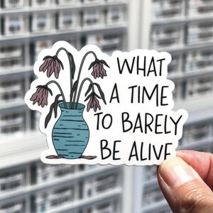 May include: A die-cut sticker featuring a blue vase with horizontal lines containing drooping, wilted purple flowers. To the right of the vase, the text reads, "WHAT A TIME TO BARELY BE ALIVE" in a bold, black, hand-lettered sans-serif font.