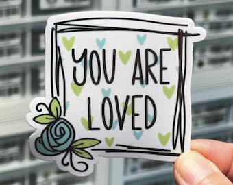 Può includere: Un adesivo rettangolare con sfondo bianco che presenta il testo nero "YOU ARE LOVED" all'interno di un bordo nero disegnato a mano. Il design include piccoli cuori azzurri e verde lime sparsi e una rosa blu con foglie verdi.