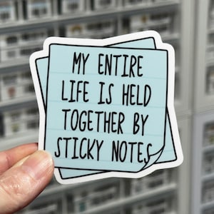 May include: A light blue sticker with the text "MY ENTIRE LIFE IS HELD TOGETHER BY STICKY NOTES" in black. The sticker is in the shape of a sticky note with a white border. The sticker is held by a hand.