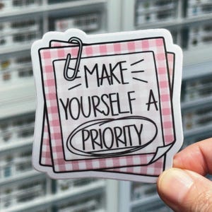 May include: A sticker with the text "MAKE YOURSELF A PRIORITY" in black, with a paperclip graphic. The sticker has a pink and white gingham pattern background. The sticker is held by a hand.