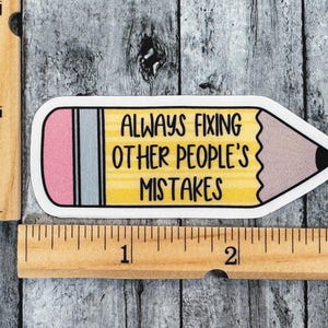 Puede incluir: Una pegatina con forma de l&aacute;piz con cuerpo amarillo, goma rosa y virola gris. El texto "ALWAYS FIXING OTHER PEOPLE'S MISTAKES" est&aacute; impreso en negro. Una regla de madera muestra que la pegatina mide aproximadamente 7,6 cm de largo.